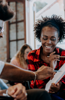 A woman smiling while looking at a tablet, sitting with a man in a casual setting with large windows, and another person in the background.