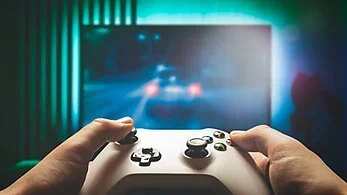 Person holding a video game controller watching a screen showing a car racing game in a dimly lit room.