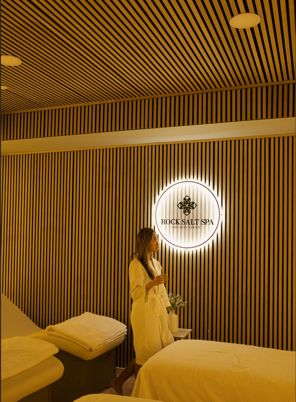 A woman in a white robe standing in a spa room, illuminated by a circular sign that reads "Rock Salt Spa Wellness Center" on a wood-panelled wall.