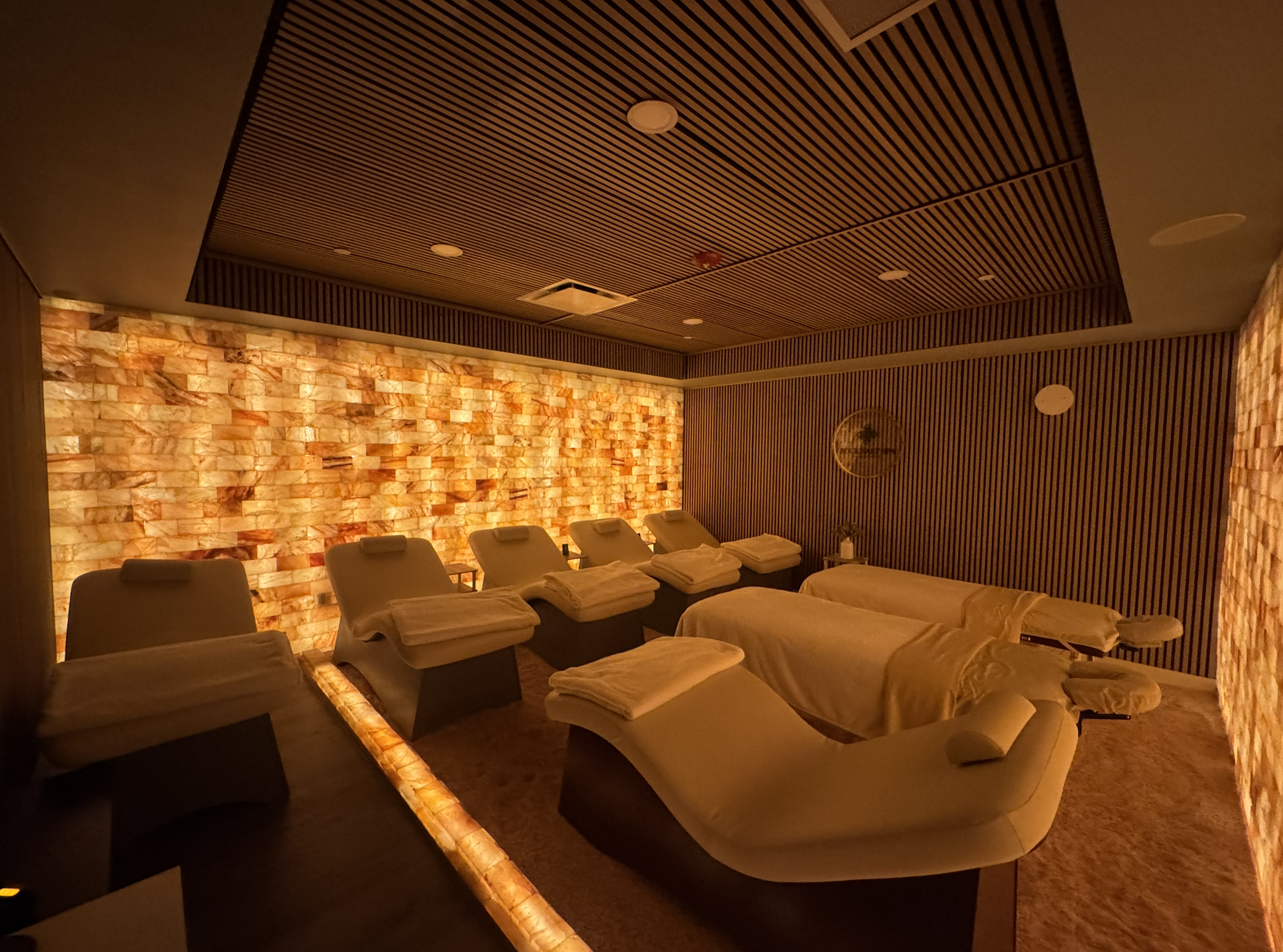 Spa treatment room with reclining chairs, rolled towels, dim lighting, salt wall, and wooden ceiling panels.