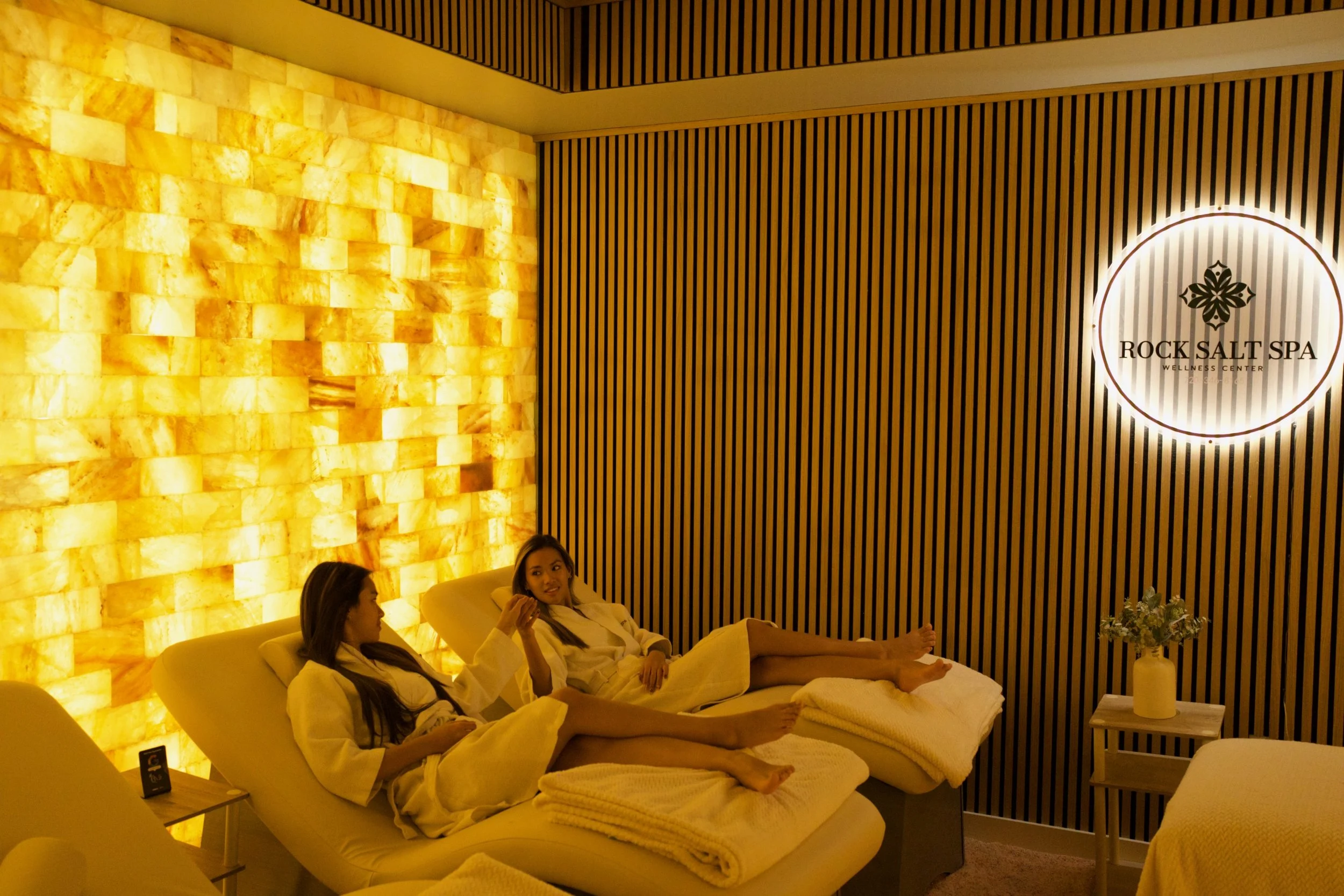 Two women relaxing on lounge chairs at Rock Salt Spa wellness center, with warm candlelit lighting and salt wall decor.