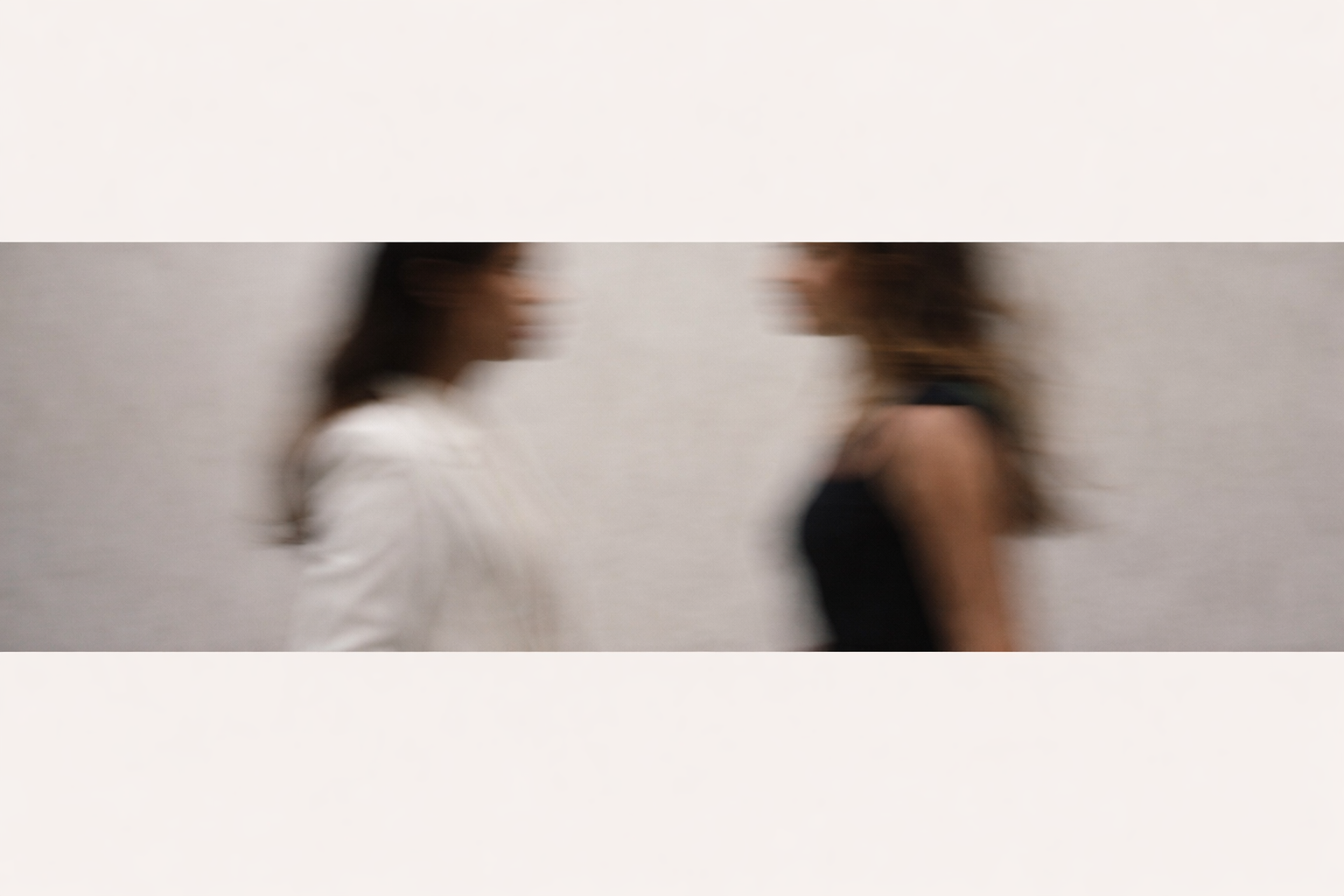 Two women walking in opposite directions, blurred due to motion, against a plain light-colored wall.