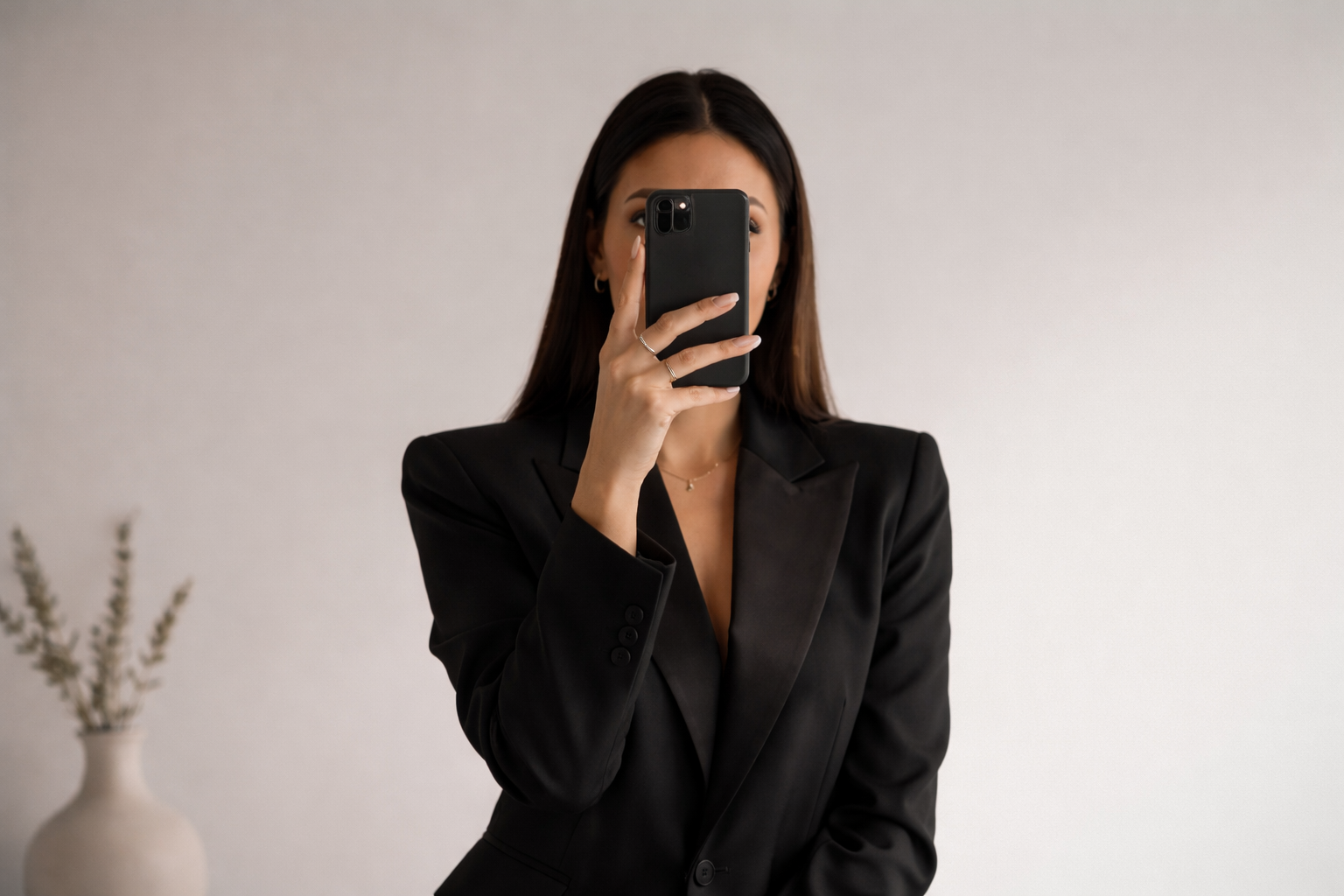 A woman in a black blazer taking a mirror selfie with a smartphone, standing in a minimalistic room with a white wall and a vase with dried plants in the background.