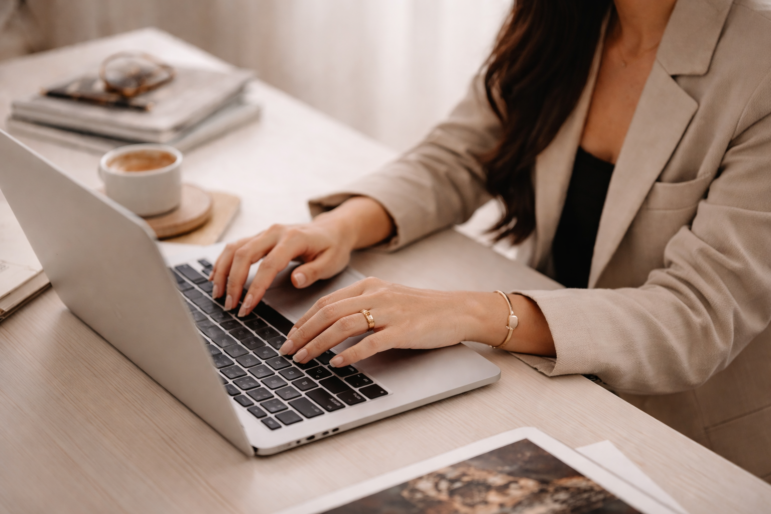 A woman using a silver laptop at a wooden desk, with a cup of coffee, a stack of books, and reading glasses nearby.