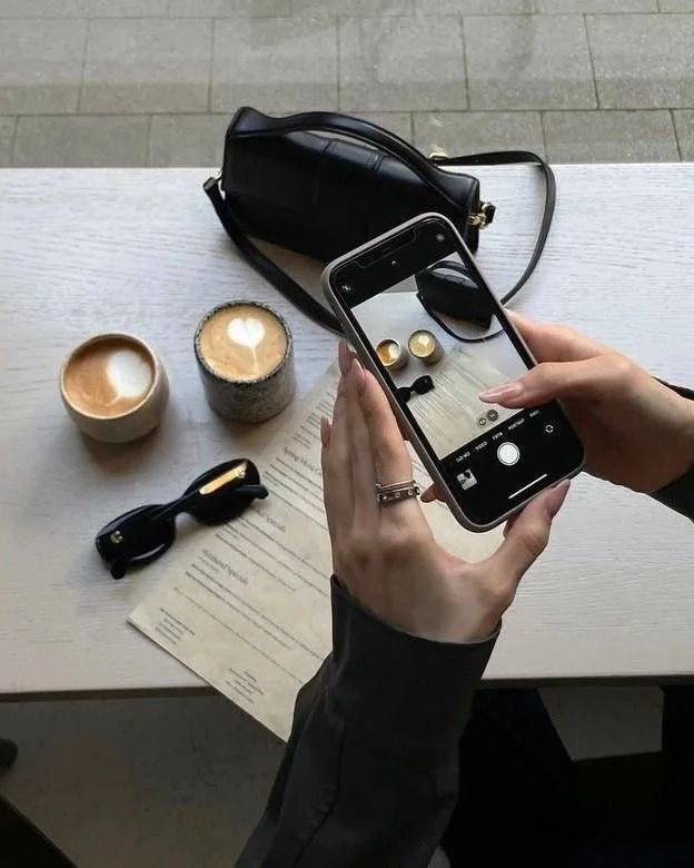 Person taking a photo of a pair of gold earrings with a smartphone at a cafe table, which also holds two cups of coffee, a black purse, sunglasses, and a piece of paper.