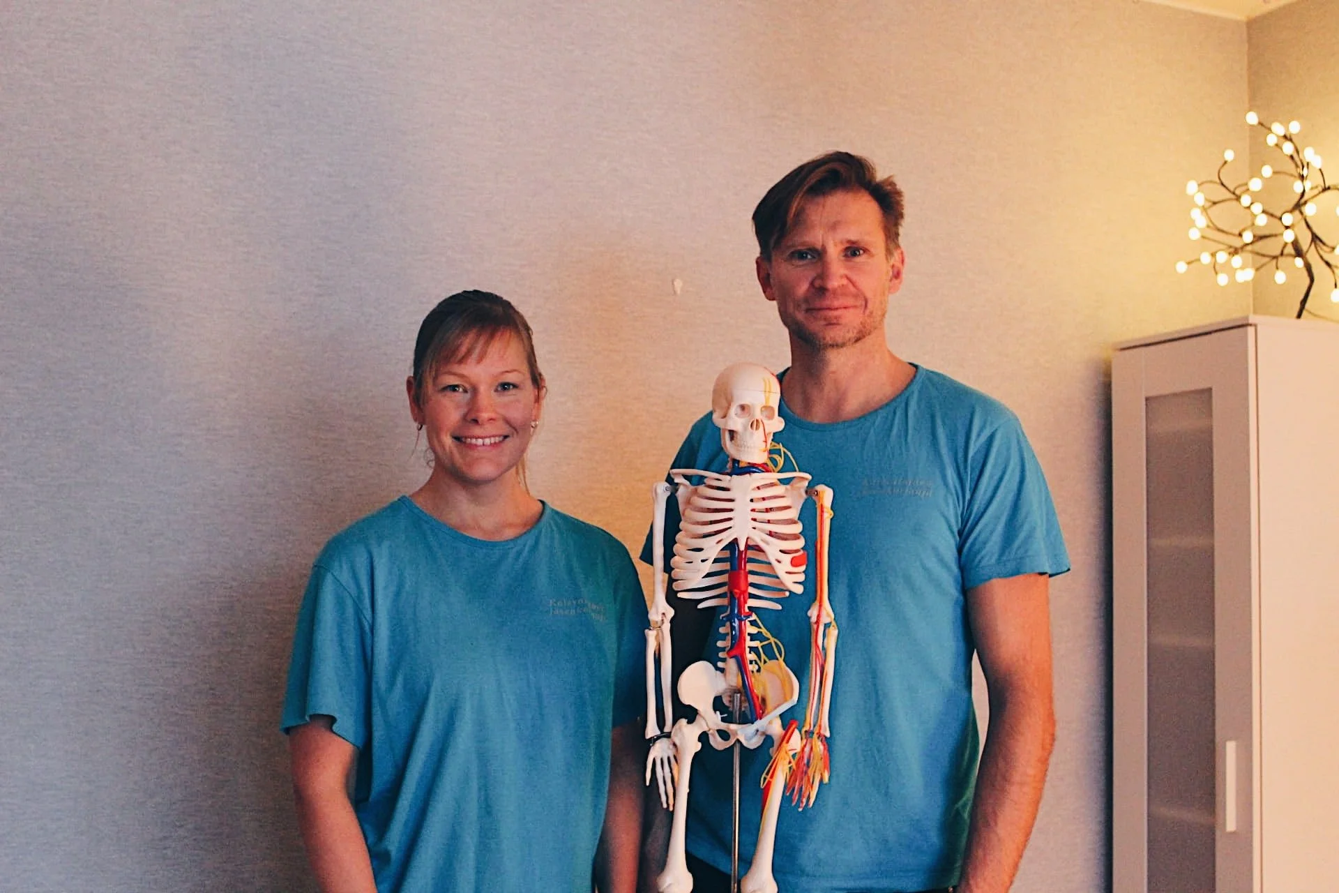 Marko ja Liisa seisovat yhdessä ja hymyilevät. Molemmat pukeutuneina sinisiin kalevalaisen jäsenkorjauksen T-paitoihin. Heidän välissään on anatominen ihmisen luuranko malli (Kalle).