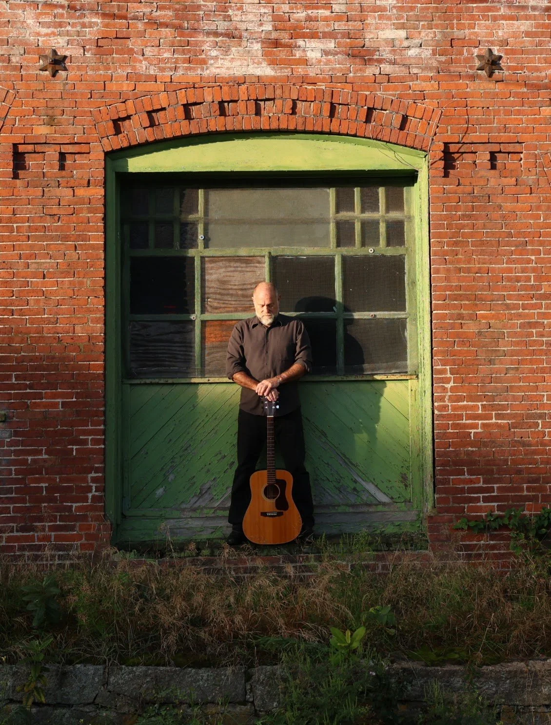 An older man with a beard stands against a green wooden door on a red brick building, holding an acoustic guitar.