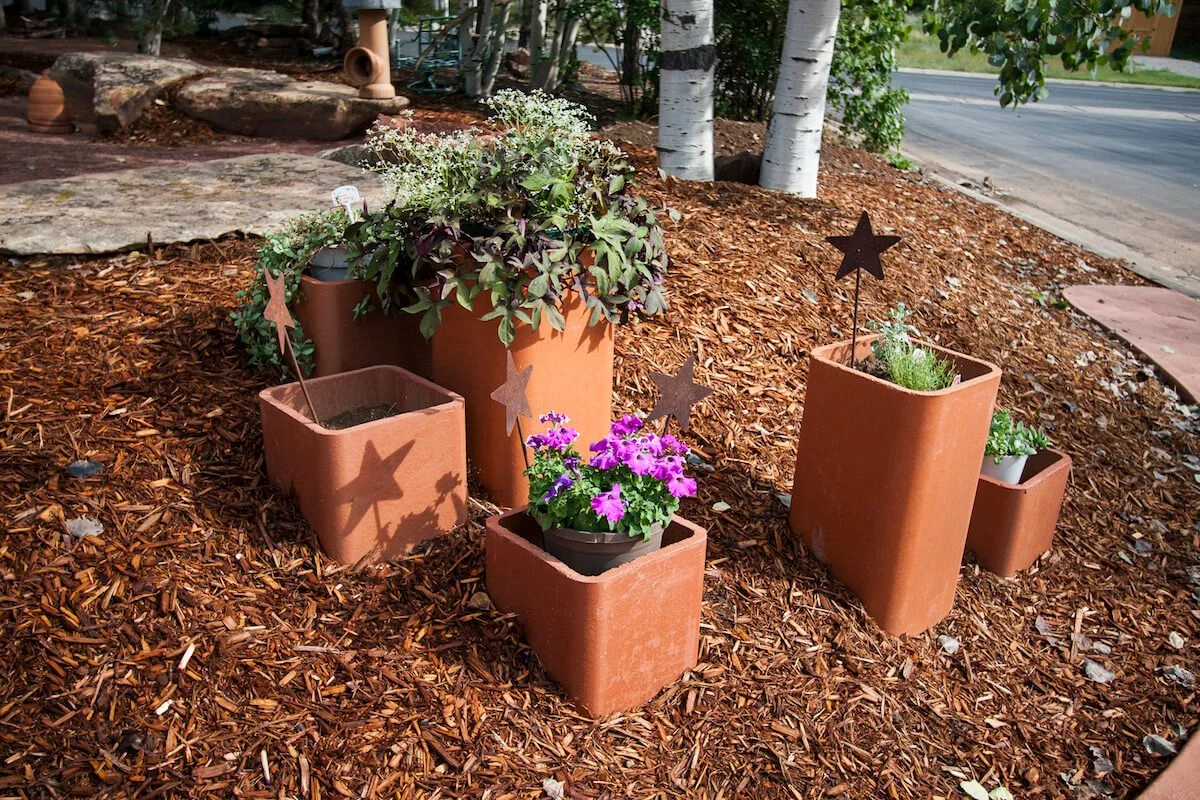 Clay pots with flowering plants and decorative star and angel stakes on a mulch bed in a garden.