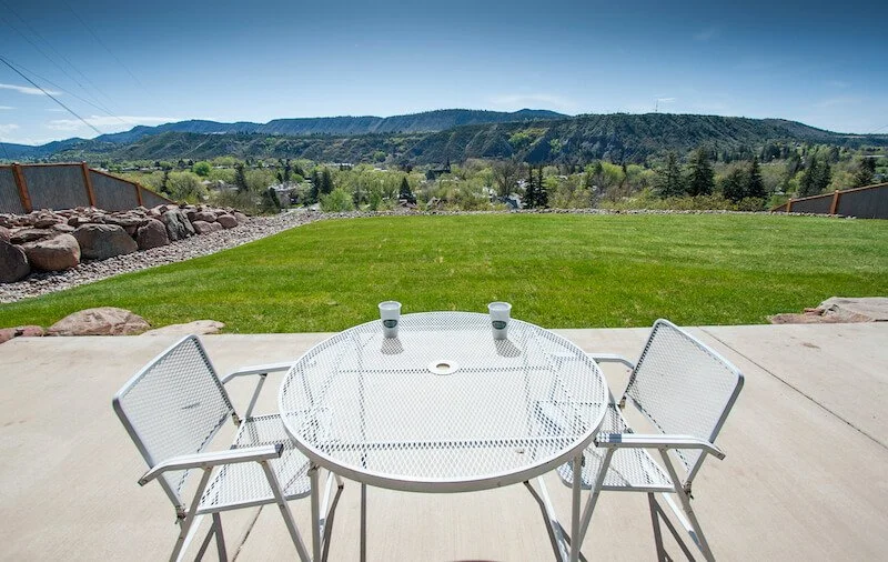 Outdoor patio with a white metal table and three white metal chairs, overlooking a backyard with green grass and a mountain landscape in the distance.