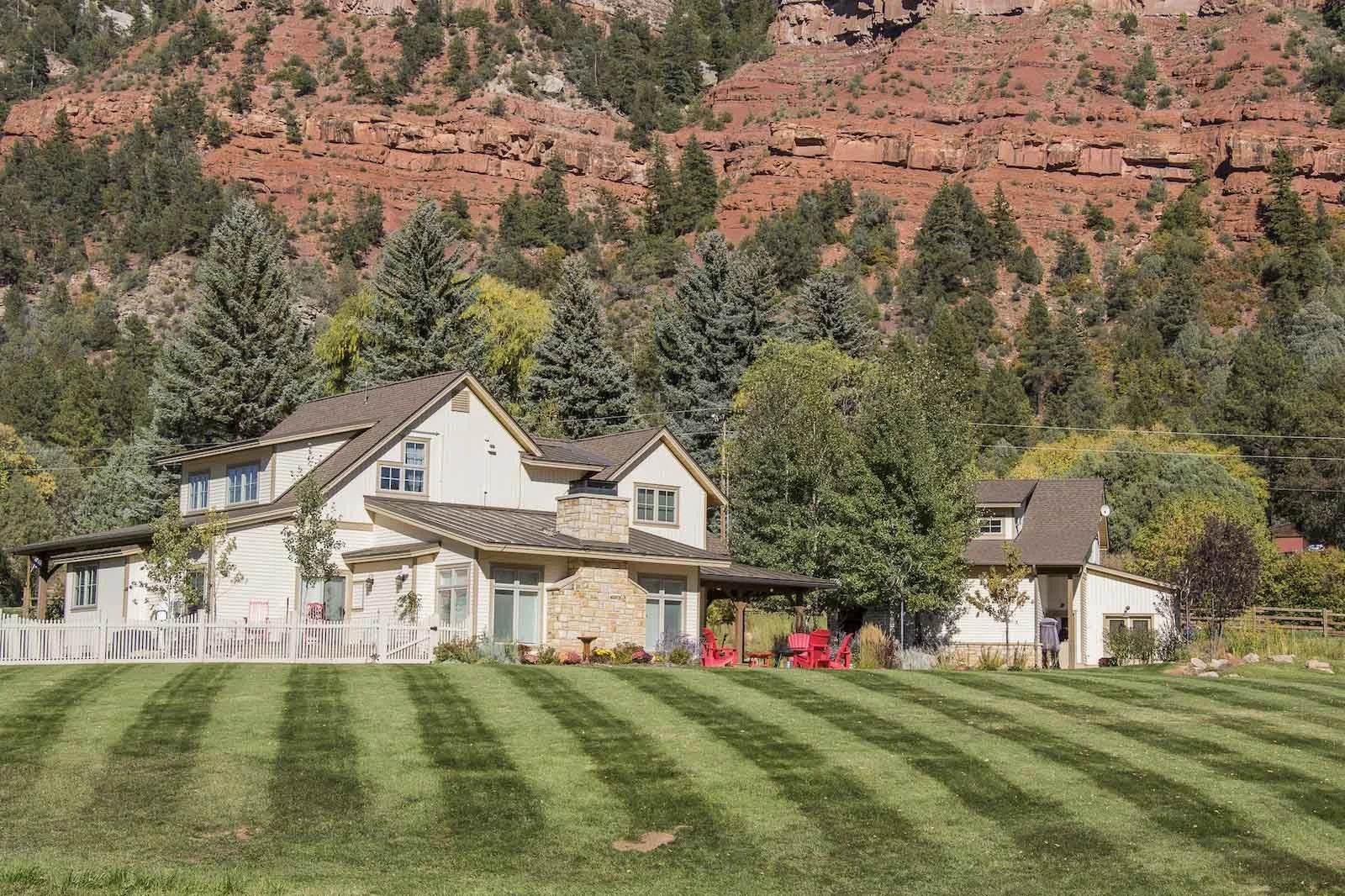 A large house with a stone chimney is situated in front of a green lawn with striped grass. There is a patio area with red chairs and a table. The background features a hillside covered with trees and reddish rock formations.