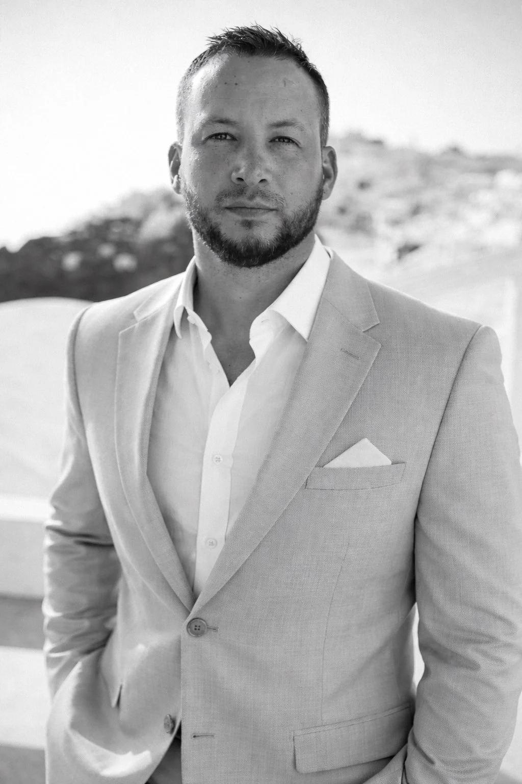 A man in a light-colored suit and white shirt standing outdoors. He has short hair and a beard, with a serious expression, looking at the camera. The background is blurred, showing a landscape.