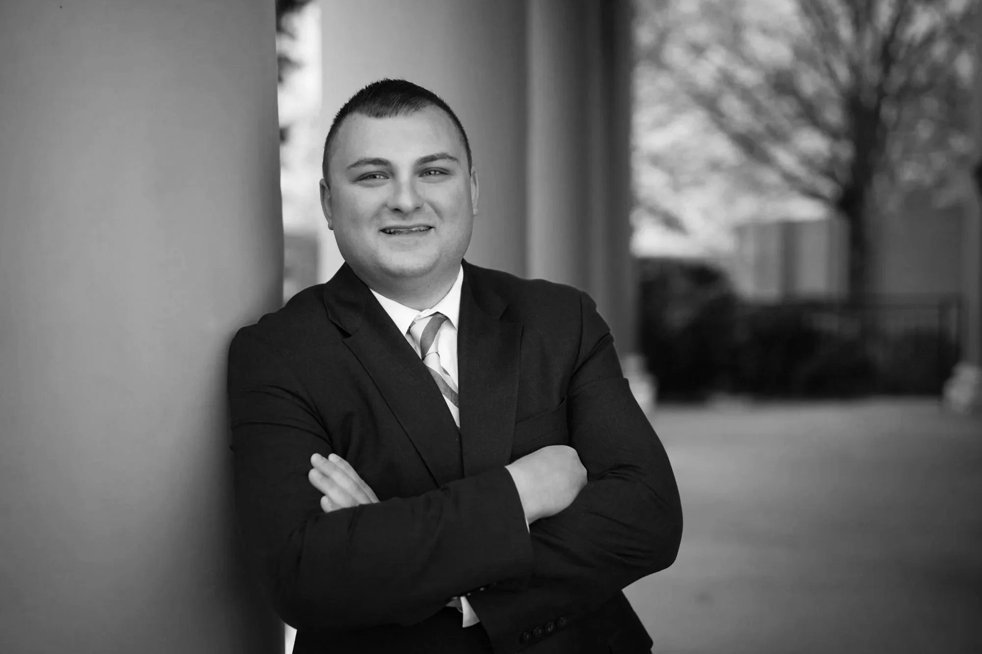 Black and white photo of a man in a suit with short hair, smiling, arms crossed, leaning against a large column outdoors with trees and a fence in the background.