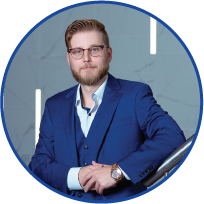 A man in a blue suit and glasses sitting at a desk in an office with modern lighting and decor.