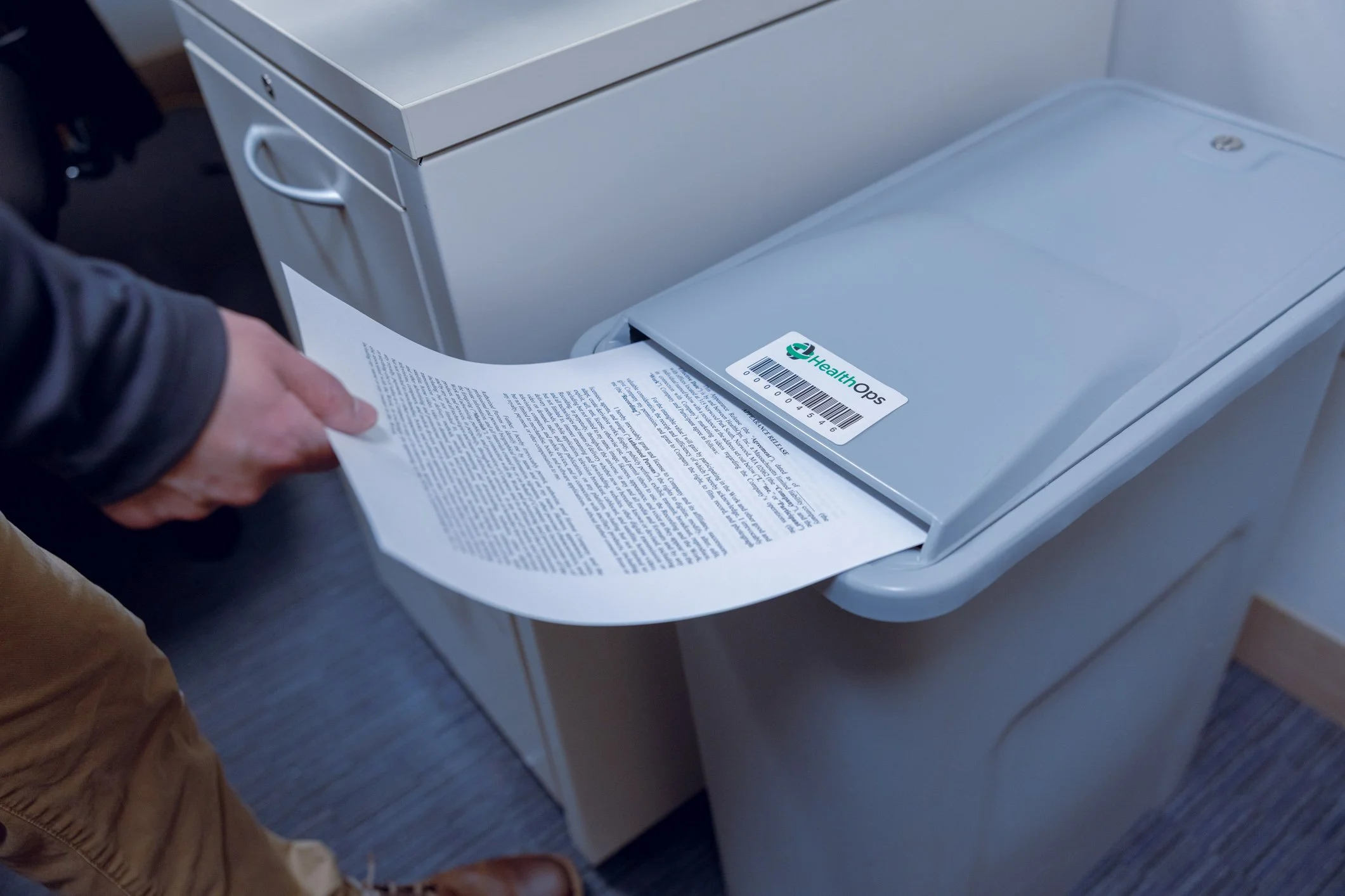 Person inserting a printed document into a gray shredder with a barcode label reading 'Health Ops'.