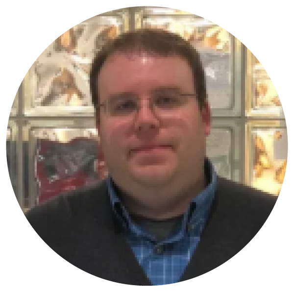 Portrait of a man with glasses, dark hair, and a slight smile, wearing a dark blazer over a blue checked shirt, in front of a display of popcorn bags.
