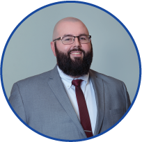 A man with glasses and a beard smiling, wearing a gray suit, white shirt, and red tie, against a light blue background