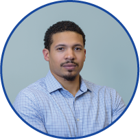 Portrait of a young man with short curly hair and a goatee, wearing a light blue checkered shirt, standing against a teal background.