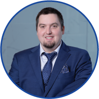 A headshot of a man in a blue suit, white shirt, and patterned tie, smiling against a light blue background.