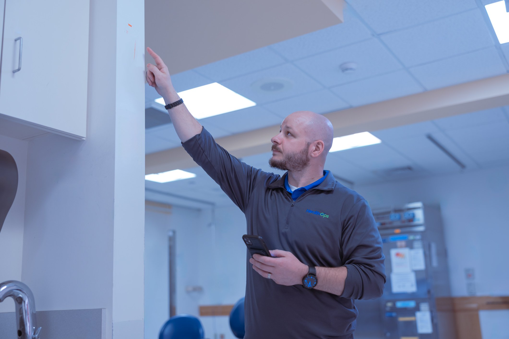 A man with a beard and a bald head, wearing a gray zip-up jacket and a black watch, is standing in a hospital corridor and pointing at a whiteboard on the wall while holding a smartphone in his other hand.