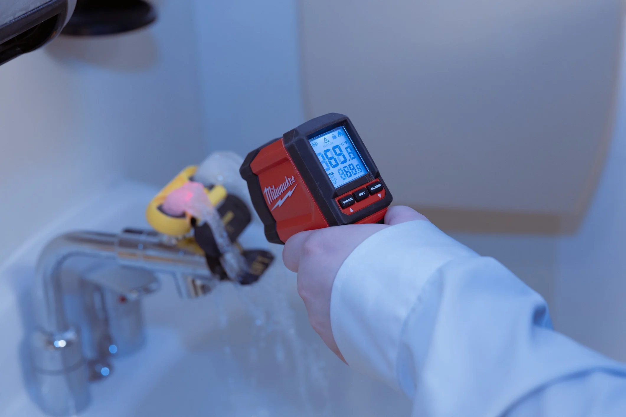 A person wearing a white lab coat holding a laser distance meter in front of a kitchen sink with a yellow and black power tool resting on top.