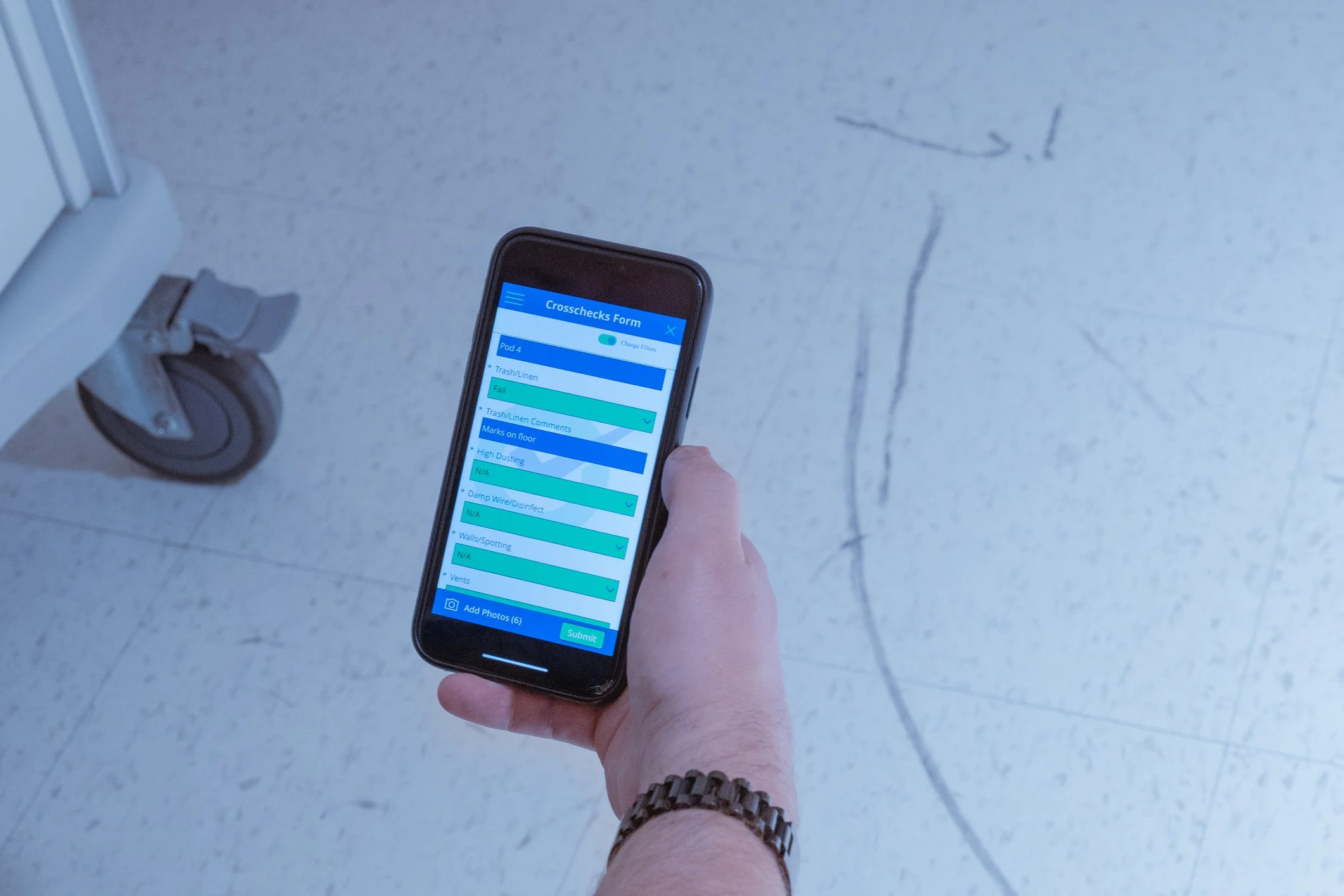 Person holding a smartphone displaying a crosscheck form, with a hospital or clinic corridor floor and a wheel of a medical cart in the background.