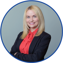 Professional woman with blonde hair wearing a dark blazer and red blouse, smiling with arms crossed, against a gray background.