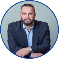 Professional man with dark hair and beard wearing a navy blazer and light blue shirt, sitting with arms crossed against a light blue background