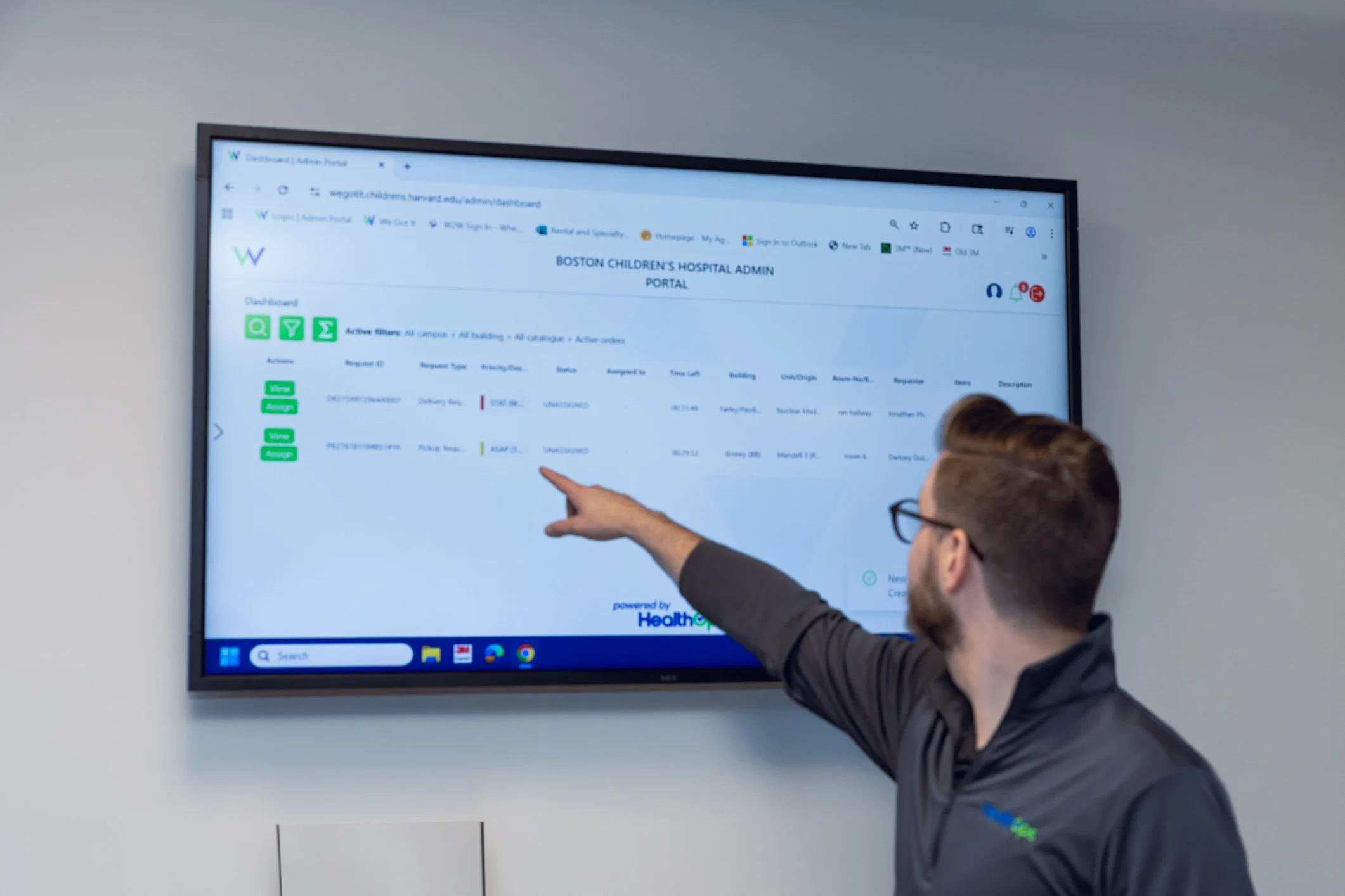 A man wearing glasses and a dark gray shirt points at a large screen displaying a hospital admin portal in a meeting room.