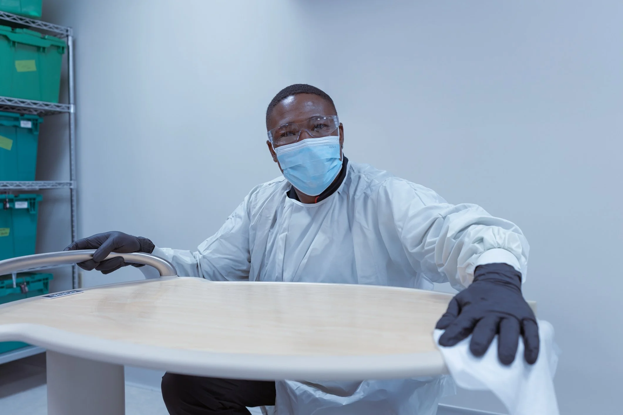 A researcher in protective gear, including a face mask, gloves, and goggles, is assembling or inspecting a piece of furniture or equipment in a laboratory or workspace.