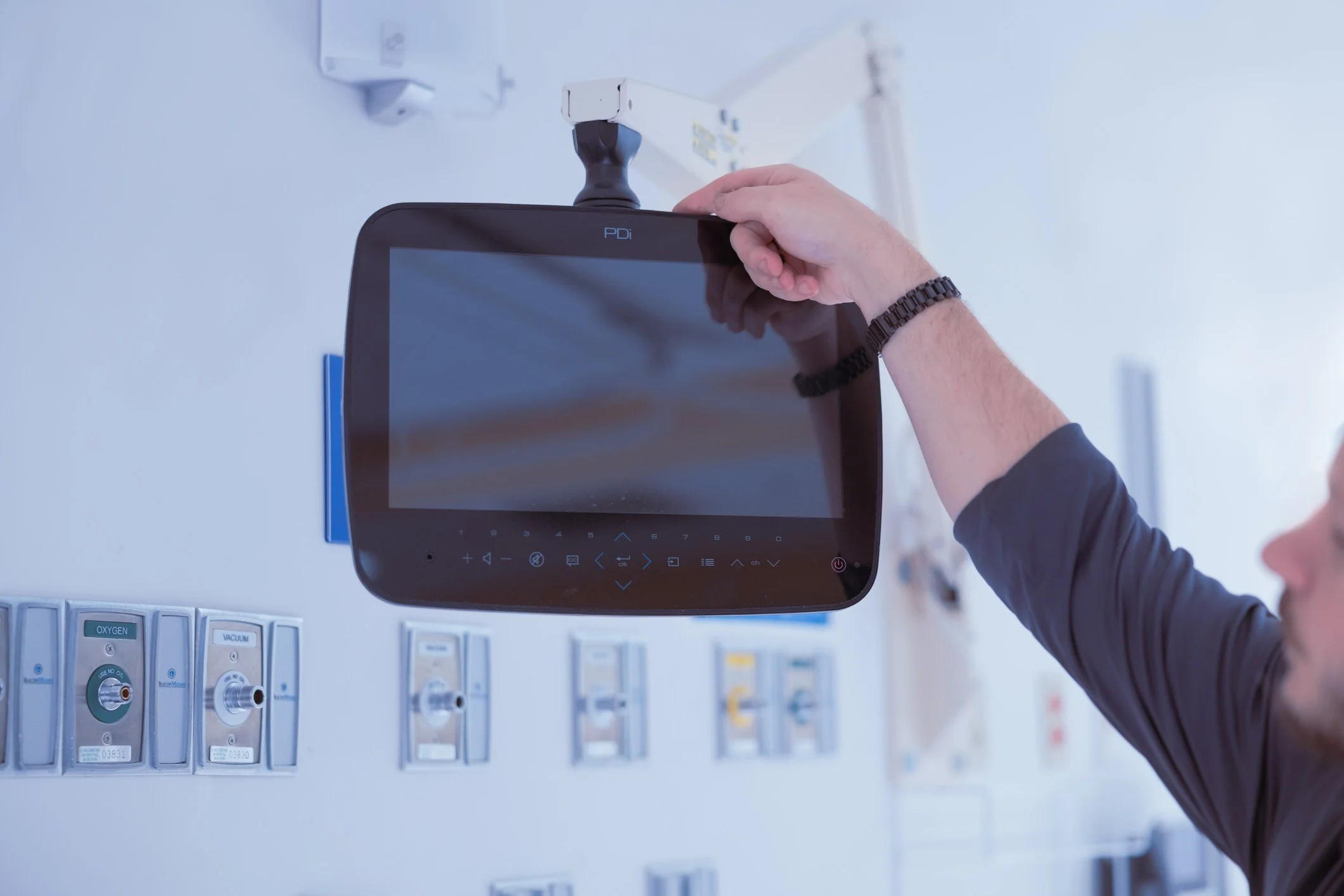 Person adjusting a medical monitor mounted on a hospital wall, with medical gas and oxygen outlets visible below.