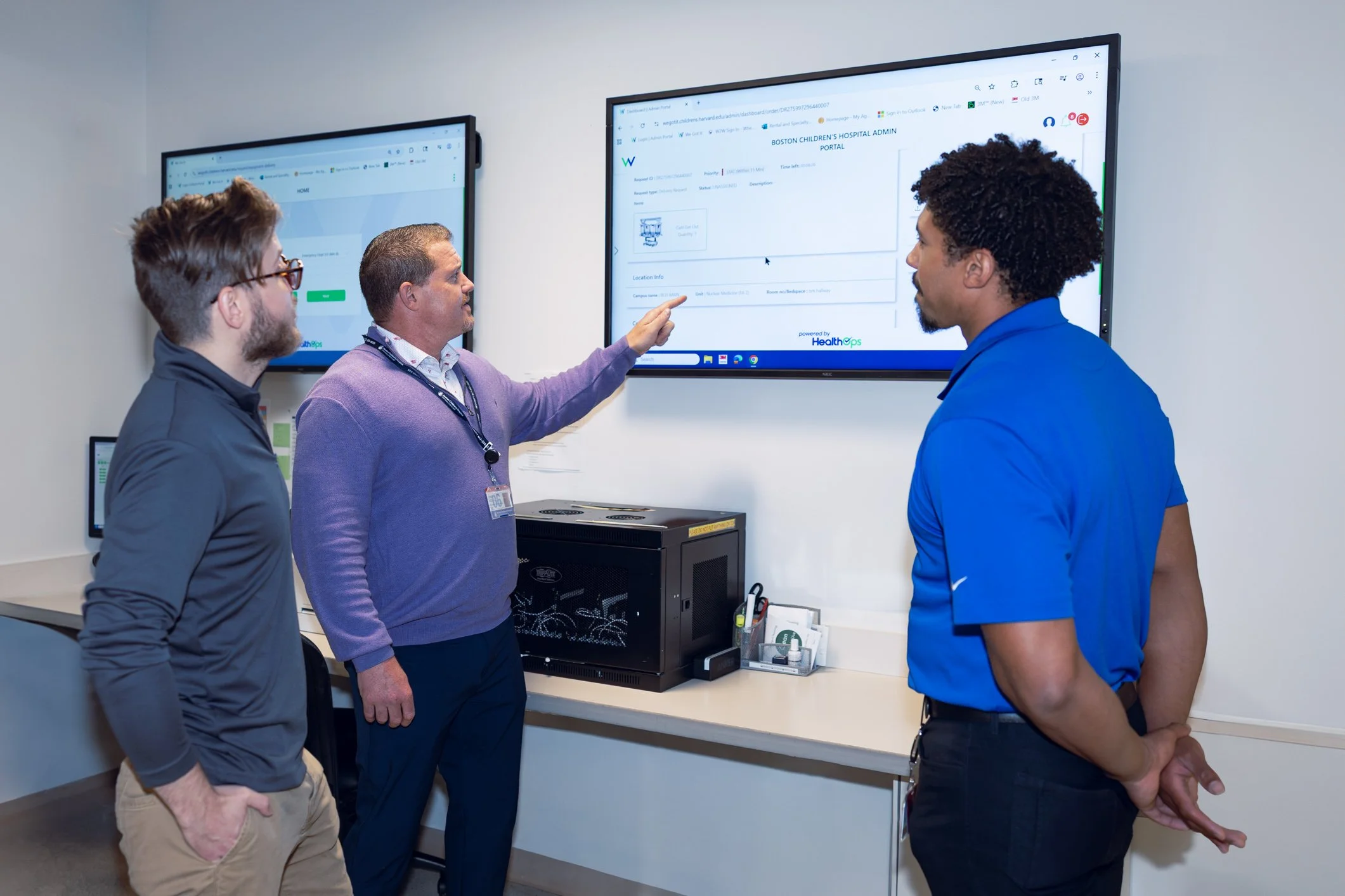 Three individuals in a medical setting are engaged in a discussion while looking at digital screens mounted on the wall. One person is pointing at the larger screen, which displays a hospital portal interface labeled 'Boston Children's Hospital Administrative Portal.' The other two individuals are listening attentively.