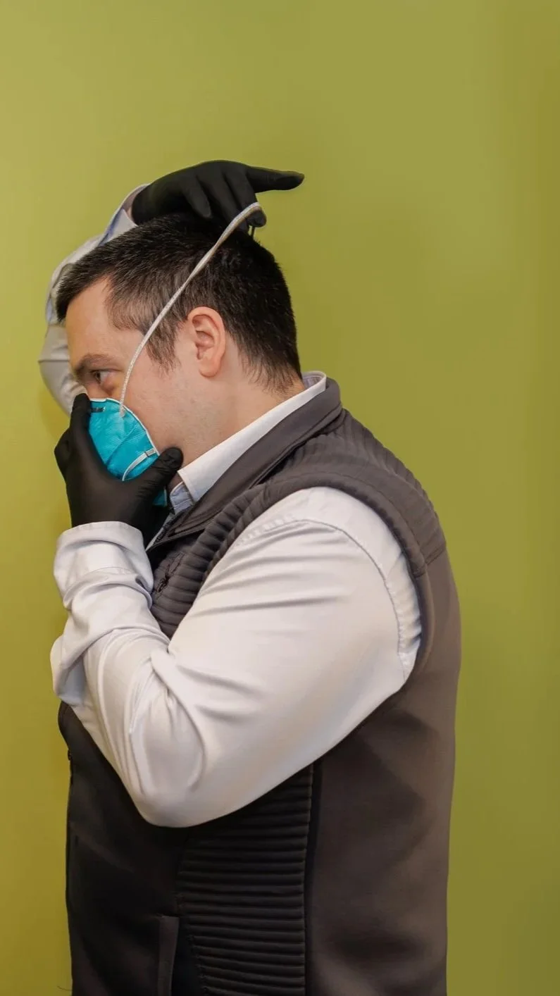 A man wearing a blue face mask and black gloves is holding his nose with one hand while using the other hand to place a pair of goggles on his head, against a green background.