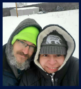 A smiling man and woman in winter clothing outdoors in a snowy landscape.