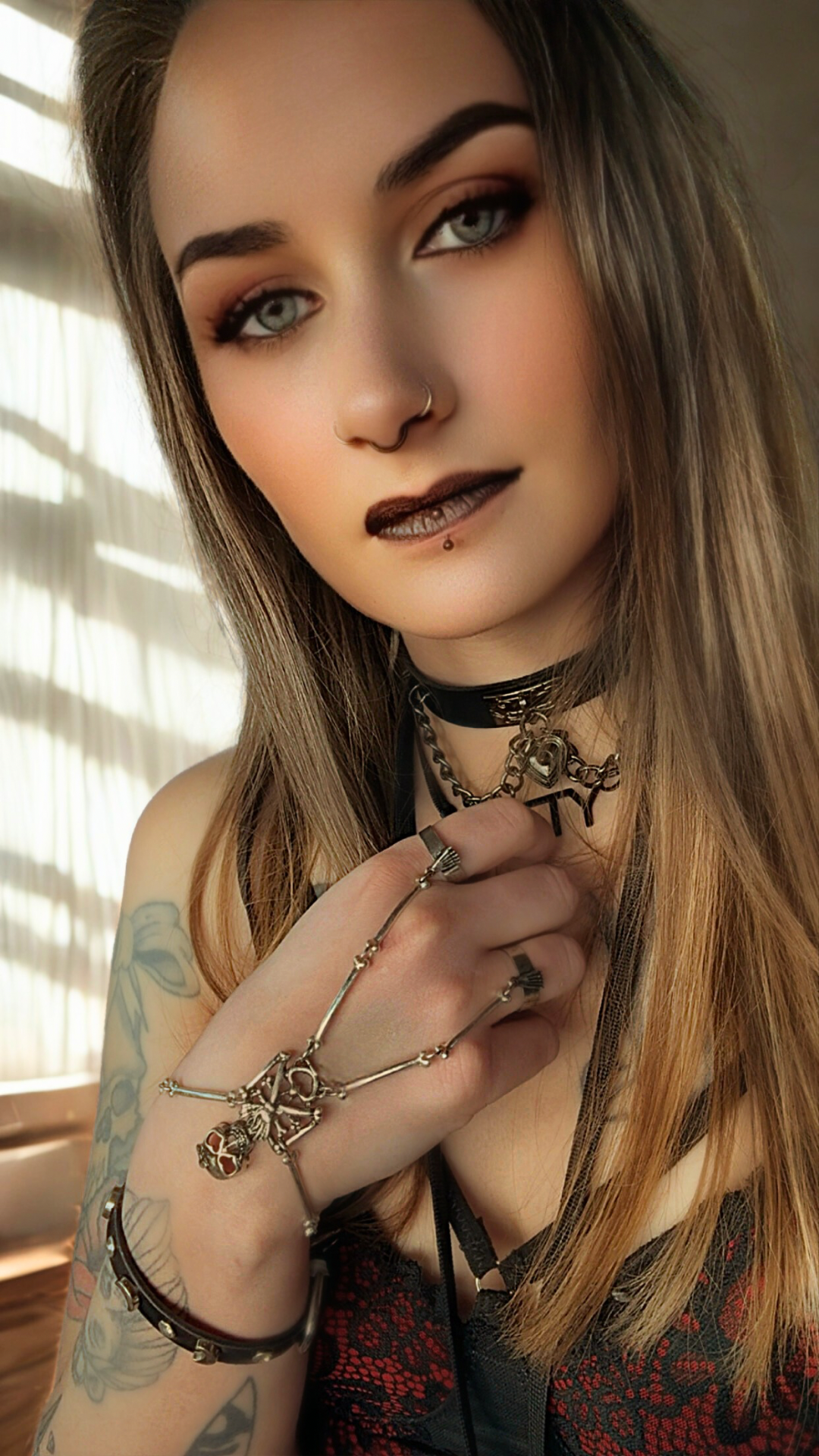 A young woman with blue eyes and dark makeup, wearing goth style jewelry including a septum ring and layered necklaces, holding her hand near her face with chrome jewelry and tattoos visible, standing in front of window blinds.