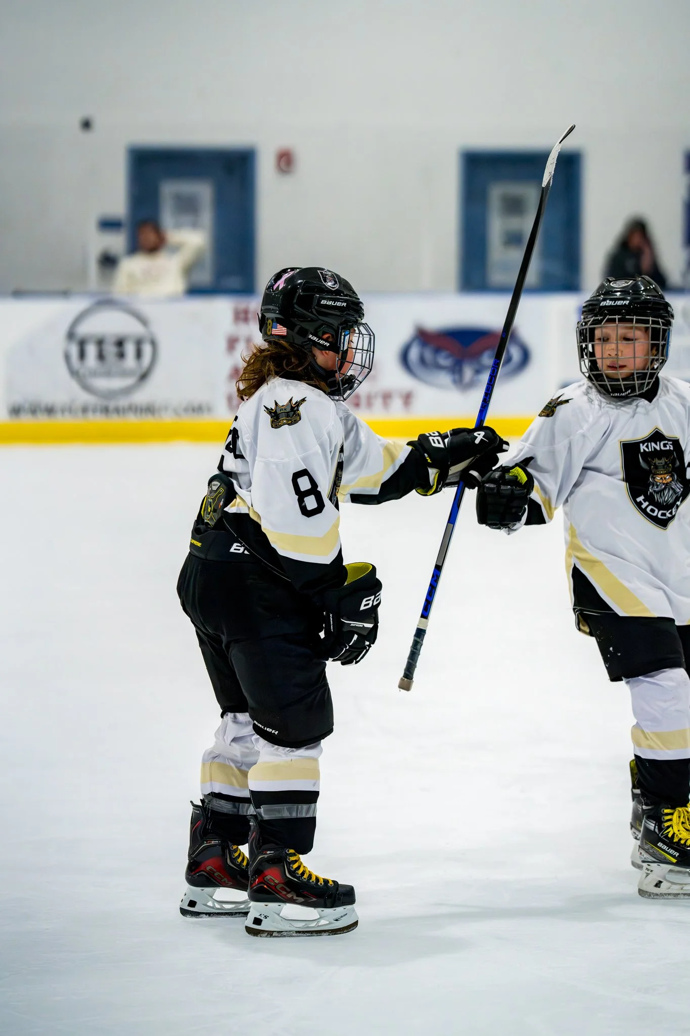 Two young hockey players in white jerseys with black and gold accents, wearing helmets and skates, are on the ice rink. They are engaged in a gesture of greeting or celebration, with one player extending her skate blade to meet the other's in a touch.