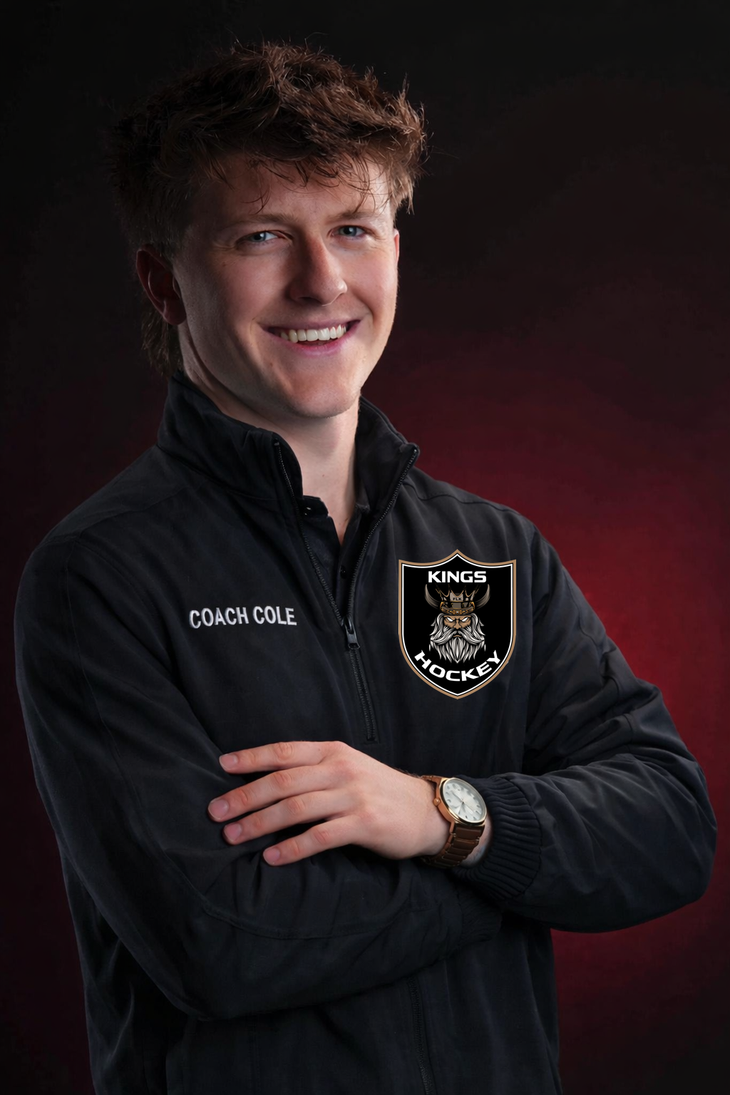 A young man in a black Coach jacket with "Coach Cole" embroidered on it, standing with arms crossed and smiling. The jacket features a logo of a Viking with the words "Kings Hockey." He is wearing a wristwatch and has a dark, blurred background.