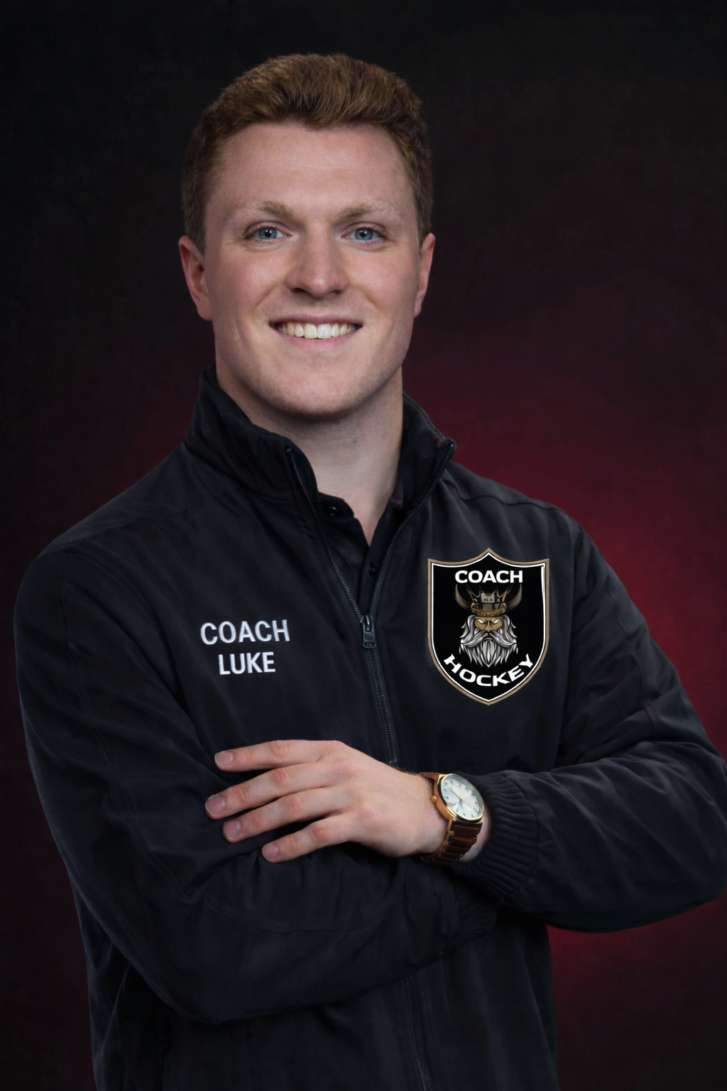 A smiling man wearing a black jacket with embroidered patches that say "Coach Luke" and a badge reading "Coach Hockey" featuring a Viking emblem. He is posing with his arms crossed, wearing a watch on his left wrist, against a dark gradient background.