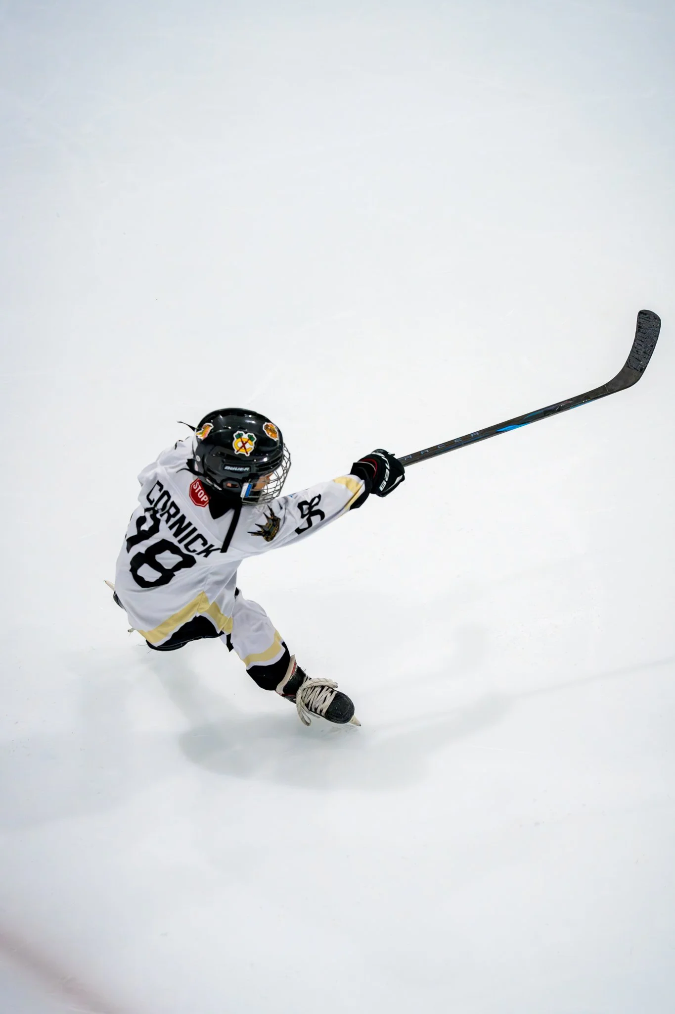A young hockey player in a white uniform with the name 'Cornick' and number 28, reaching out with a hockey stick on an ice rink.