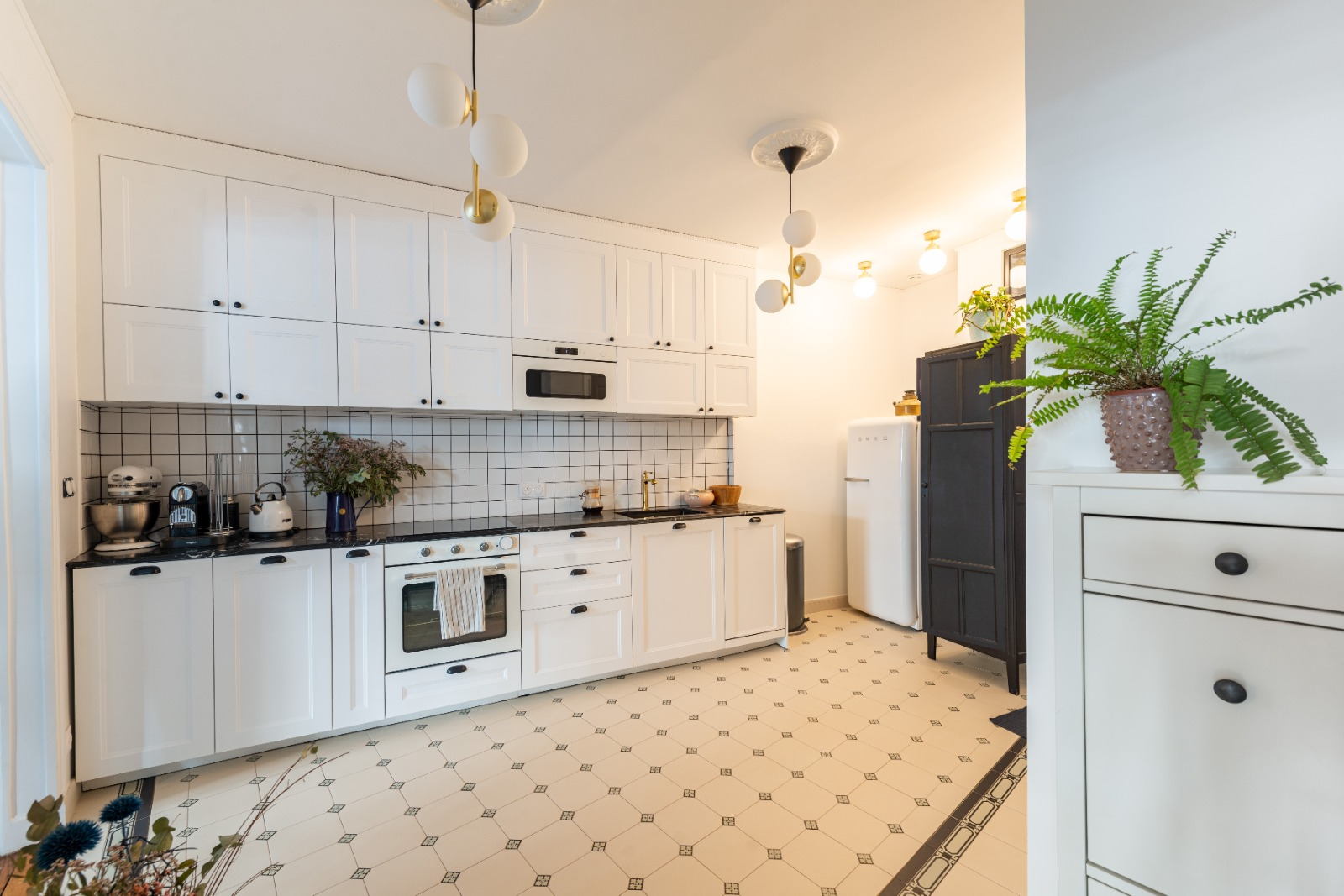 Kitchen with white cabinets, black countertop, white tiled backsplash, vintage black and white patterned floor, kitchen appliances, plants, and modern lighting fixtures.