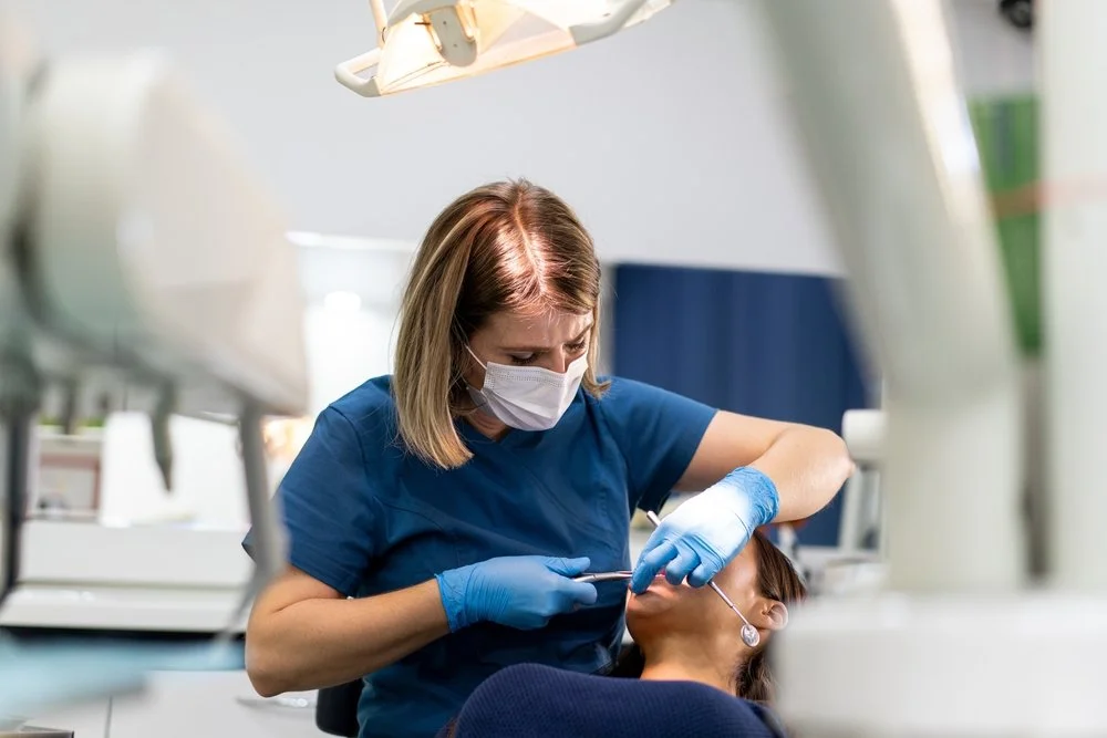 A female healthcare professional in blue scrubs and a face mask providing dental care to a patient lying in a dental chair, with dental equipment around.