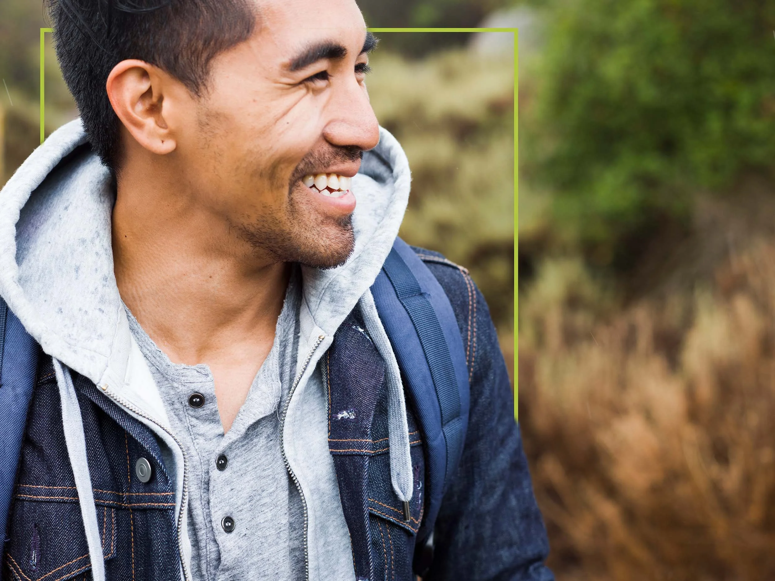 A man outdoors, smiling, wearing a hoodie and denim jacket, with a backpack, in a natural setting with blurred greenery and plants in the background.