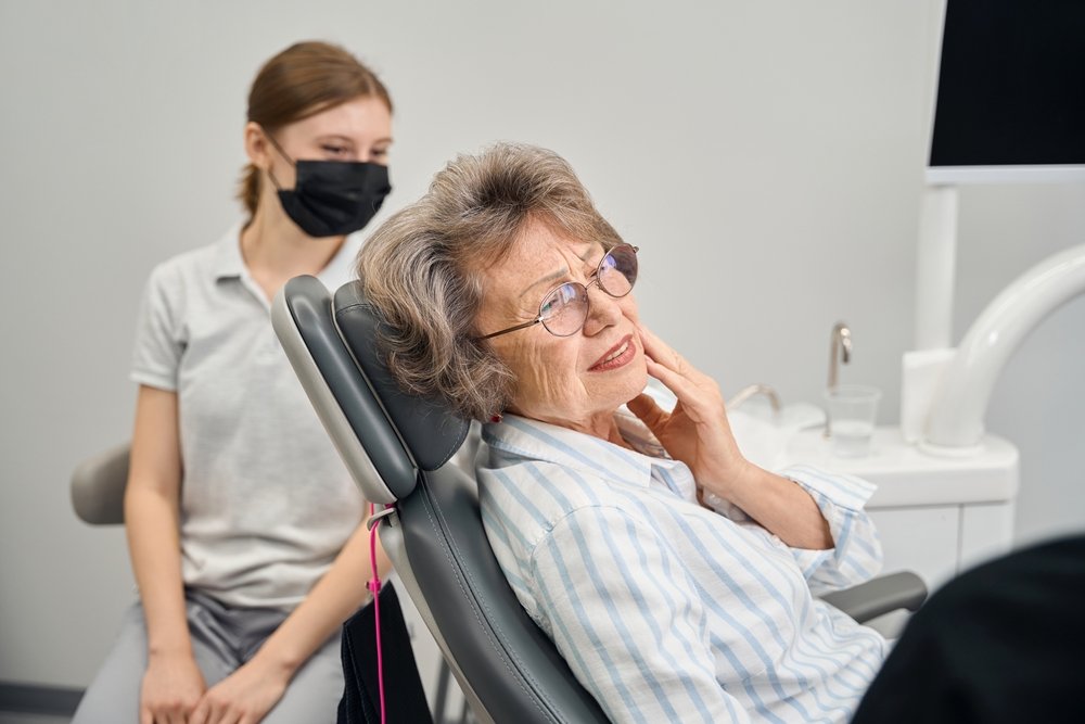 An elderly woman in a dental chair, smiling with her hand on her cheek, while a dental assistant wearing a black face mask stands nearby in a dental office.