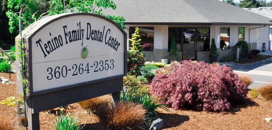 Exterior view of Tenino Family Dental Center with a sign in front and a building with large windows behind, surrounded by landscaped plants and bushes.