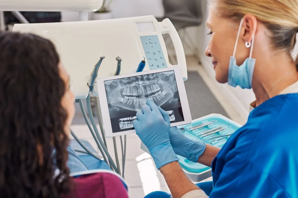 Dentist examining a dental X-ray on a tablet with a patient, using dental tools nearby.