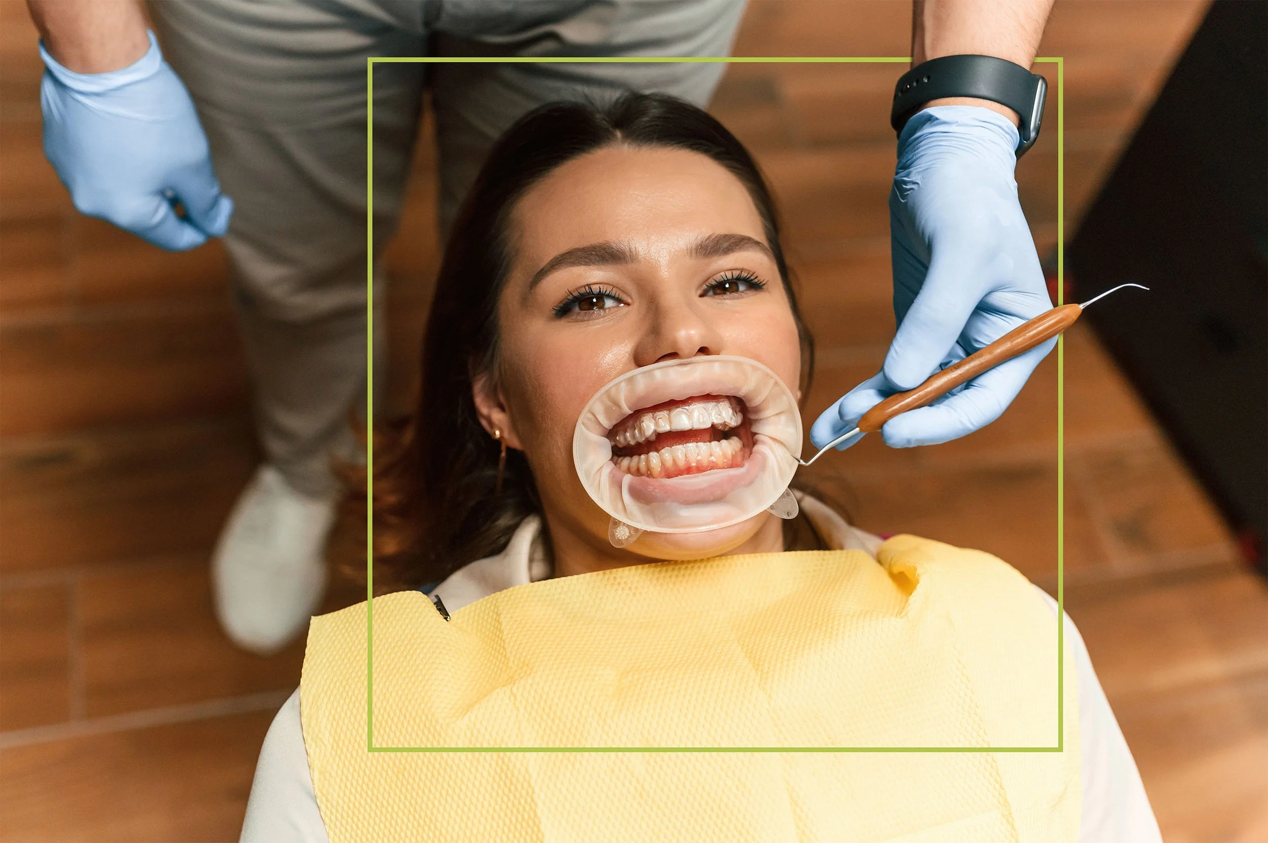 A woman lying down in a dental chair with her mouth open, wearing a rubber dental mouth gag showing her teeth, while a dentist in gloves uses a dental tool.