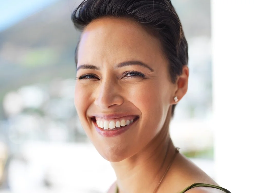 A smiling woman with short dark hair, wearing pearl earrings and a necklace, outdoors during daytime.
