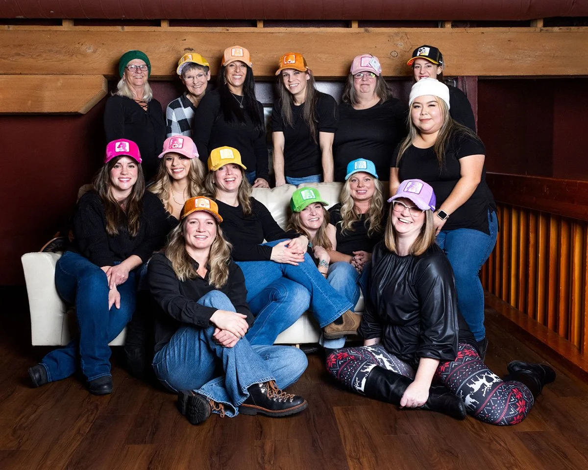 Group of thirteen women indoors, wearing colorful hats, some with holiday or festive themed clothing, sitting and standing on a couch and floor, smiling for a group photo.