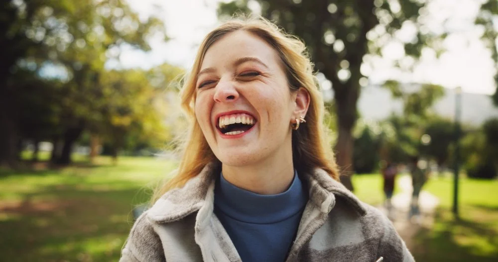 A woman with blonde hair laughing outdoors in a park with trees in the background.
