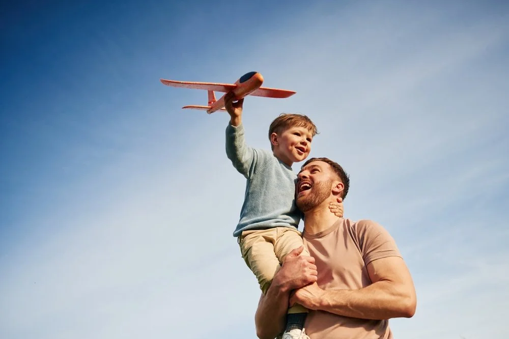 A man holding a young boy on his shoulder, with the boy holding a toy airplane up in the air.