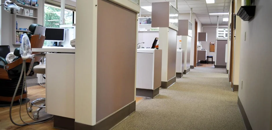 Medical clinic reception area with a work desk, a computer monitor, and a dental chair on the left, separated by beige partitions. The right side has a hallway with multiple cubicles and chairs, leading to another room at the back.